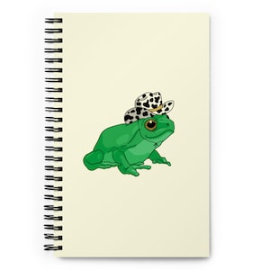 May include: A green frog wearing a black and white spotted cowboy hat. The frog is sitting on a light yellow background.