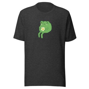 May include: A dark heather grey t-shirt with a cartoon frog holding a white flower. The frog is green with a light green belly and has a happy expression.