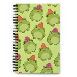 Puede incluir: Una libreta espiral verde con un patrón de ranas verdes que llevan fresas rojas y frambuesas moradas sobre un fondo verde claro.