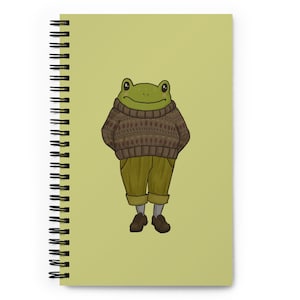 Op de afbeelding: Een groene kikker die een bruin en wit gepatroneerd trui en een groene broek draagt. De kikker staat op een lichtgroene achtergrond.