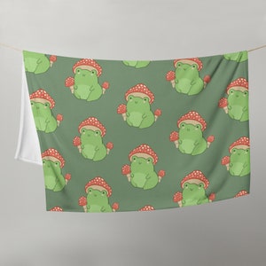 Peut inclure: Une tapisserie en tissu vert avec un motif répété de grenouilles vertes tenant des champignons rouges et blancs. Le fond est de couleur vert foncé.