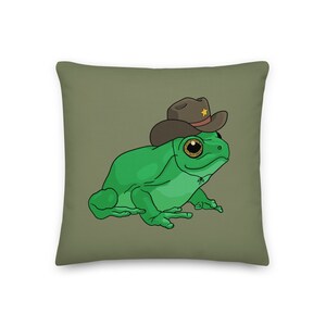 Puede incluir: Cojín cuadrado con una ilustración de dibujos animados de una rana verde con un sombrero de vaquero marrón con una estrella dorada. El cojín tiene un fondo verde oliva liso.