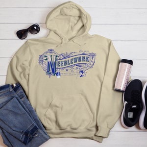 May include: Beige hooded sweatshirt with a blue and white vintage logo that reads "Needlework Society Est. 1920" with decorative flourishes. The logo includes the text "Only the finest silks and linens"