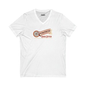 Op de afbeelding: Wit V-hals T-shirt met een afbeelding met de tekst "Introvert maar bereid om kruissteken te bespreken". Het ontwerp heeft een cirkelvormig embleem en een banner in bruin- en roodtinten. Het shirt is gemaakt van een zacht, comfortabel materiaal.