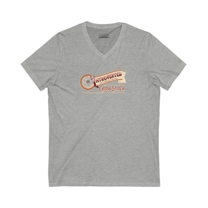 Op de afbeelding: Een heather grijze V-hals t-shirt met de tekst "INTROVERTED but willing to discuss CROSS STITCH" in een banner ontwerp. De banner is in tinten van bruin, rood en beige. Het shirt is gemaakt van een zacht materiaal.