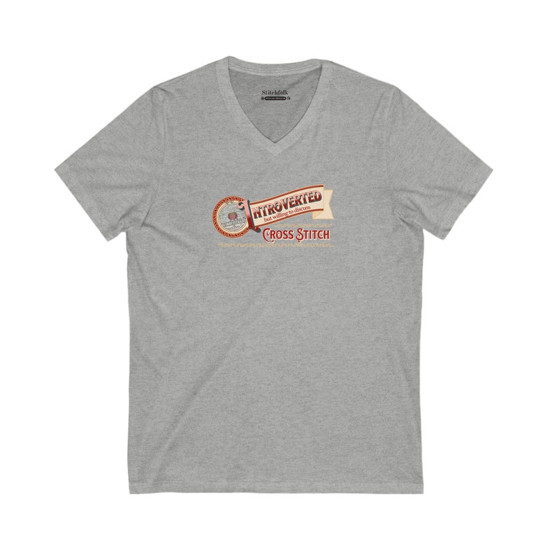 Op de afbeelding: Een heather grijze V-hals t-shirt met de tekst "INTROVERTED but willing to discuss CROSS STITCH" in een banner ontwerp. De banner is in tinten van bruin, rood en beige. Het shirt is gemaakt van een zacht materiaal.