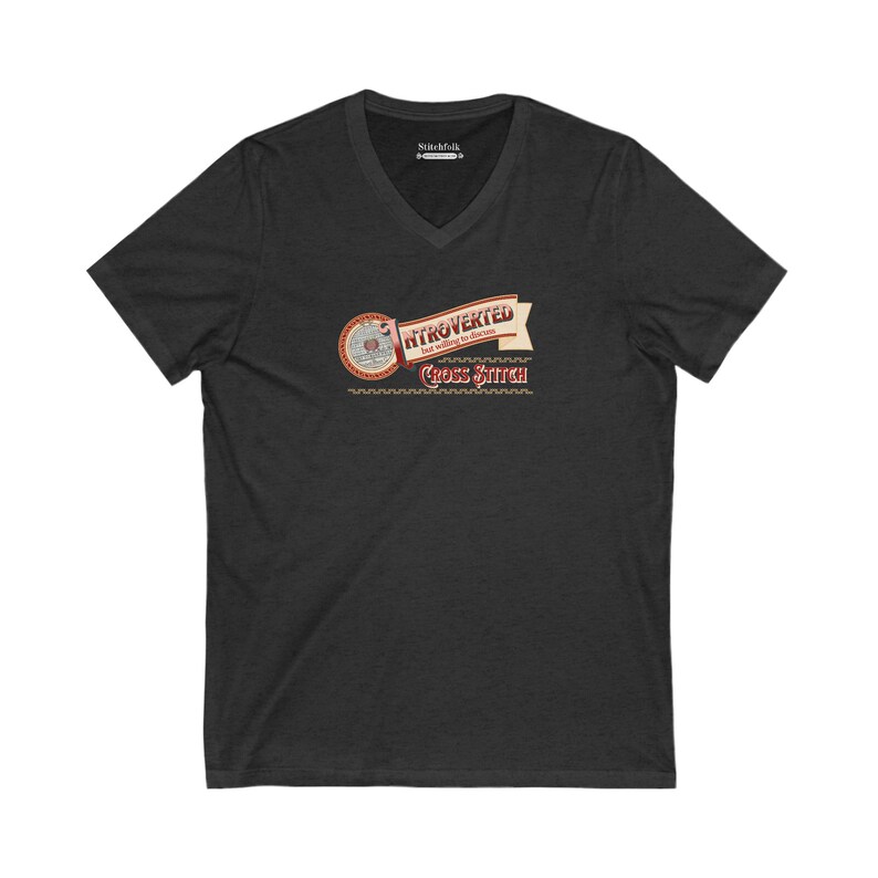 Op de afbeelding: Donkergrijs V-hals T-shirt met de tekst "Introverted but willing to discuss Cross Stitch" in een vintage design. De afbeelding bevat een cirkelvormig element met naald en draad, en een banner met het woord "Introverted".