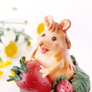 Può includere: Una piccola figurina di argilla marrone di un topo che tiene una fragola rossa. Il topo ha il naso rosa e la lingua che sporge. Il topo è circondato da foglie verdi e ci sono altre due fragole sullo sfondo.