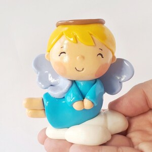 May include: A small, blue angel figurine with a halo, yellow hair, and wings. The angel is sitting on a white cloud.
