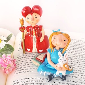 May include: A red and white Queen of Hearts figurine with a crown and a scepter, and a blue Alice in Wonderland figurine holding a white rabbit. Both figurines are made of ceramic and are sitting on an open book.