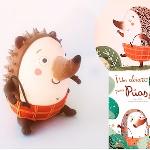 May include: A brown and white hedgehog figurine with a red and orange plaid skirt. The figurine is holding a brown basket. The image also shows a book cover with a similar hedgehog illustration and the text "&iexcl;Un abrazo para P&iacute;as!"
