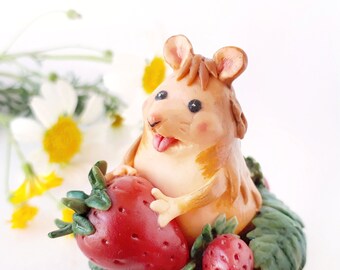 May include: A small, brown, clay figurine of a mouse holding a red strawberry. The mouse has a pink nose and tongue sticking out. The mouse is surrounded by green leaves and there are two more strawberries in the background.