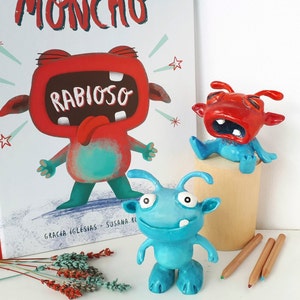 May include: Three handmade clay monster figurines. One red monster with a big mouth and the text "Rabioso" on its chest. One blue monster with a big smile and two antennas. One red monster with a big mouth and two antennas. The monsters are sitting on a wooden surface.