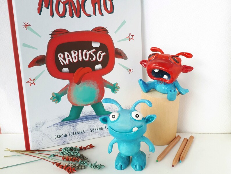 May include: Three handmade clay monster figurines. One red monster with a big mouth and the text "Rabioso" on its chest. One blue monster with a big smile and two antennas. One red monster with a big mouth and two antennas. The monsters are sitting on a wooden surface.