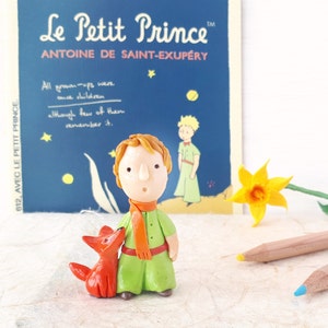 May include: A handmade figurine of The Little Prince from the book by Antoine de Saint-Exup&eacute;ry. The Prince is wearing a green tunic, orange scarf, and brown boots. He is standing next to his fox friend, who is orange and has a white belly. The figurine is in front of a book cover with the title "Le Petit Prince" and the author's name.