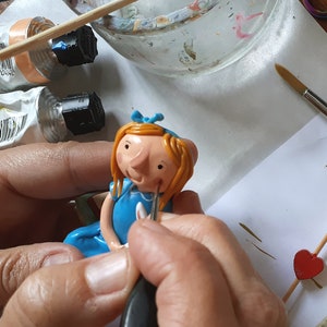 May include: A person is painting a small clay doll with a blue dress and blonde hair. The doll is wearing a blue bow and has a painted face. The person is using a small brush to paint the doll's face.