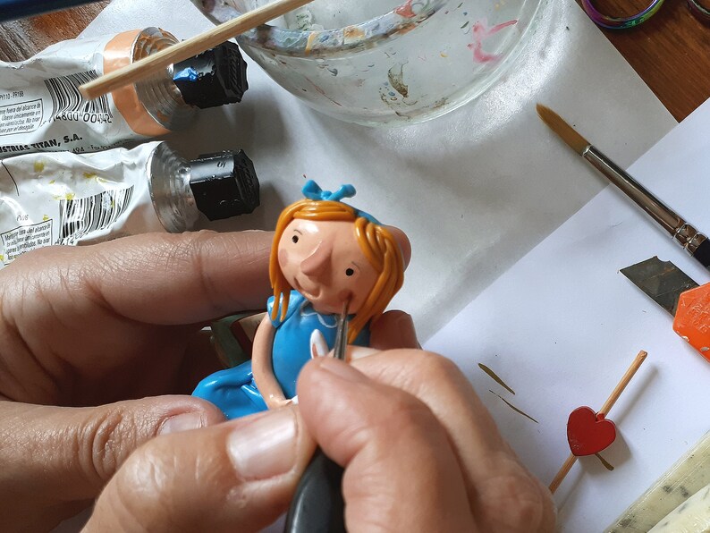 May include: A person is painting a small clay doll with a blue dress and blonde hair. The doll is wearing a blue bow and has a painted face. The person is using a small brush to paint the doll's face.