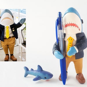May include: A blue shark figurine wearing a blue jacket, yellow tie, and brown trousers, holding a blue pen. The figurine is standing on a white surface. A small blue shark figurine is in the foreground.
