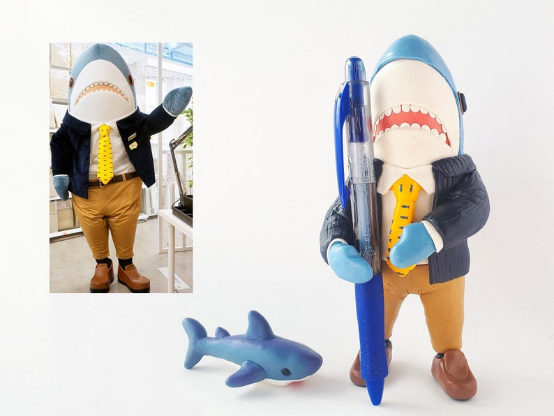 May include: A blue shark figurine wearing a blue jacket, yellow tie, and brown trousers, holding a blue pen. The figurine is standing on a white surface. A small blue shark figurine is in the foreground.