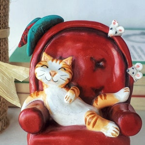 May include: A ceramic figurine of a cat relaxing in a red armchair with two white mice on the armrests. The cat has orange and white fur and a happy expression.