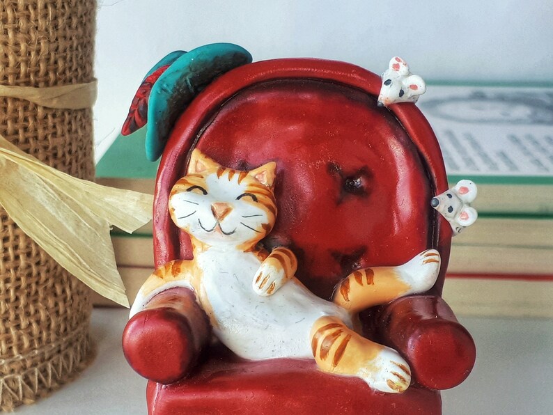 May include: A ceramic figurine of a cat relaxing in a red armchair with two white mice on the armrests. The cat has orange and white fur and a happy expression.