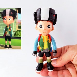 May include: A hand-crafted figurine of a cartoon character wearing a black and white helmet, a green and yellow jacket, blue trousers, and black shoes. The figurine is made of polymer clay.