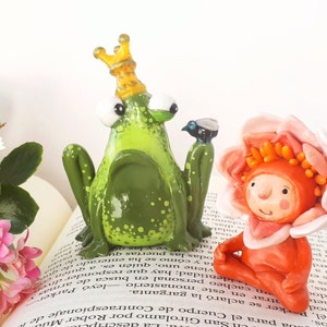 May include: A green frog wearing a gold crown sits next to a pink flower with a red face and orange hair. Both figures are made of clay and are sitting on a book.