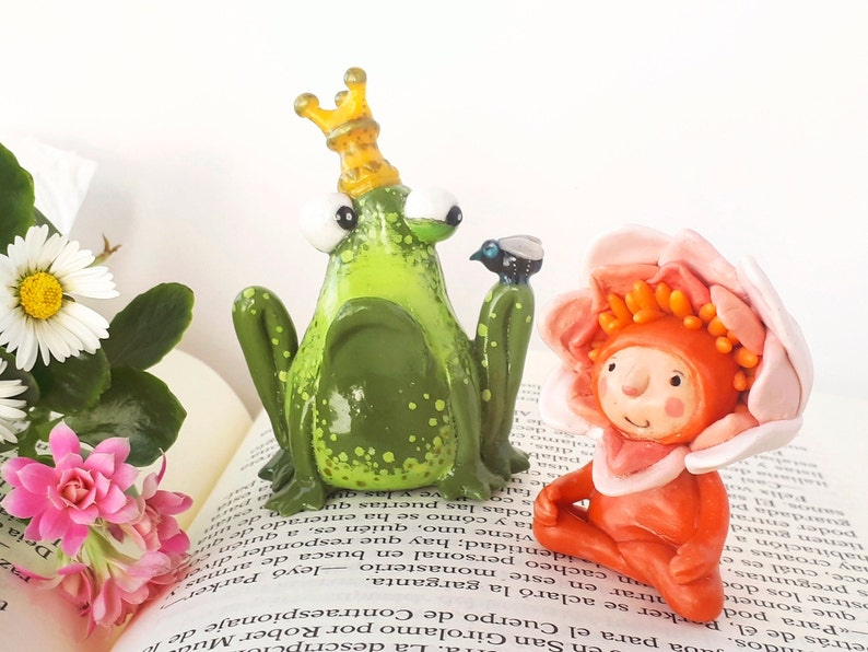 May include: A green frog wearing a gold crown sits next to a pink flower with a red face and orange hair. Both figures are made of clay and are sitting on a book.