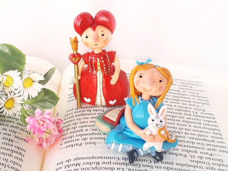 May include: A red and white Queen of Hearts figurine with a crown and a scepter, and a blue Alice in Wonderland figurine holding a white rabbit. Both figurines are made of ceramic and are sitting on an open book.