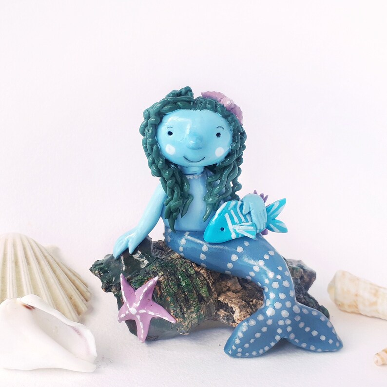 May include: A blue mermaid figurine with green hair and a blue and white spotted tail, holding a blue fish. The mermaid is sitting on a rock with a pink starfish.