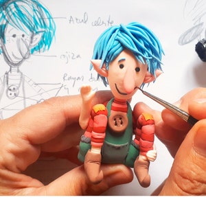 May include: A blue-haired, red and green-clothed, clay figurine with a large nose and a friendly smile. The figurine is being painted with a small brush. The image text reads "Best Wishes Creatures" and "CUSTOM FIGURES FROM DRAWINGS & PHOTOS".