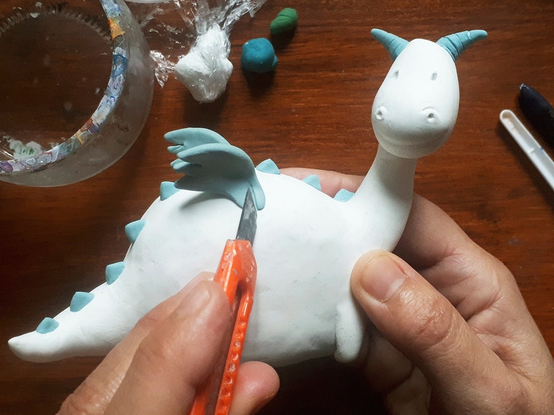 May include: A white ceramic dragon figurine with blue wings and spikes being sculpted with a craft knife. The dragon has a simple, cartoon-like face.