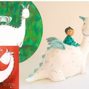 May include: A white ceramic dragon figurine with a child riding on its back. The dragon has blue wings and a green and white polka dot shirt. The book cover in the background reads "El Dragon Blanco".