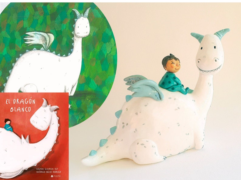 May include: A white ceramic dragon figurine with a child riding on its back. The dragon has blue wings and a green and white polka dot shirt. The book cover in the background reads "El Dragon Blanco".