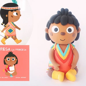 May include: A handmade clay figurine of a girl wearing a colorful dress and a headband with feathers. The figurine is sitting with her legs crossed and her hands in her lap. The figurine is titled "TERESA LA PRINCESA" and is by the authors "MAGDALENA DEL MAZO" and "CECILIA MORENO".