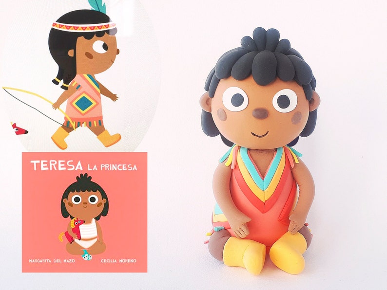 May include: A handmade clay figurine of a girl wearing a colorful dress and a headband with feathers. The figurine is sitting with her legs crossed and her hands in her lap. The figurine is titled "TERESA LA PRINCESA" and is by the authors "MAGDALENA DEL MAZO" and "CECILIA MORENO".