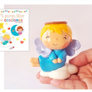 May include: A small blue angel figurine with a halo, wings, and a cloud base. The angel has a smiling face and is holding its hands together in prayer. The figurine is made of polymer clay and is hand-painted. The text on the book in the background reads "Mi primer libro de oraciones con todas".