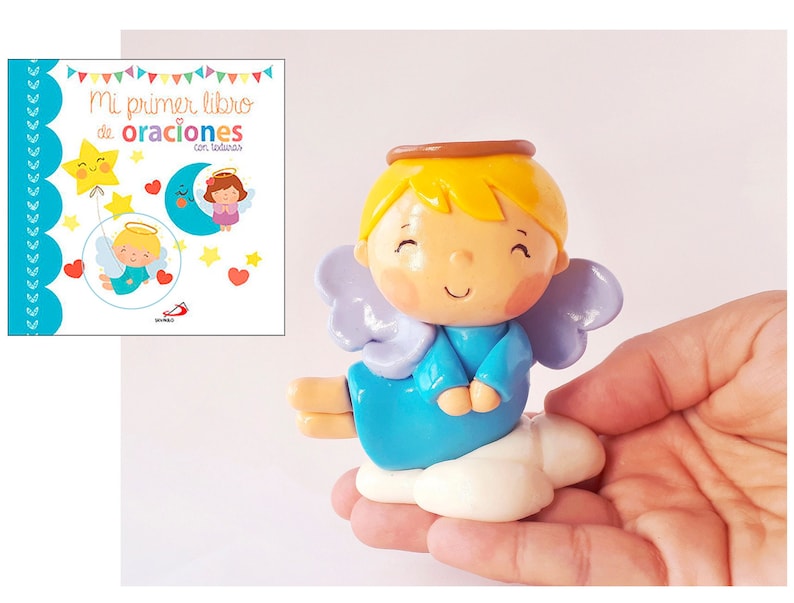 May include: A small blue angel figurine with a halo, wings, and a cloud base. The angel has a smiling face and is holding its hands together in prayer. The figurine is made of polymer clay and is hand-painted. The text on the book in the background reads "Mi primer libro de oraciones con todas".