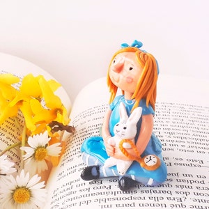May include: A blue and white porcelain figurine of Alice from Alice in Wonderland holding a white rabbit. The figurine is sitting on an open book with yellow flowers.