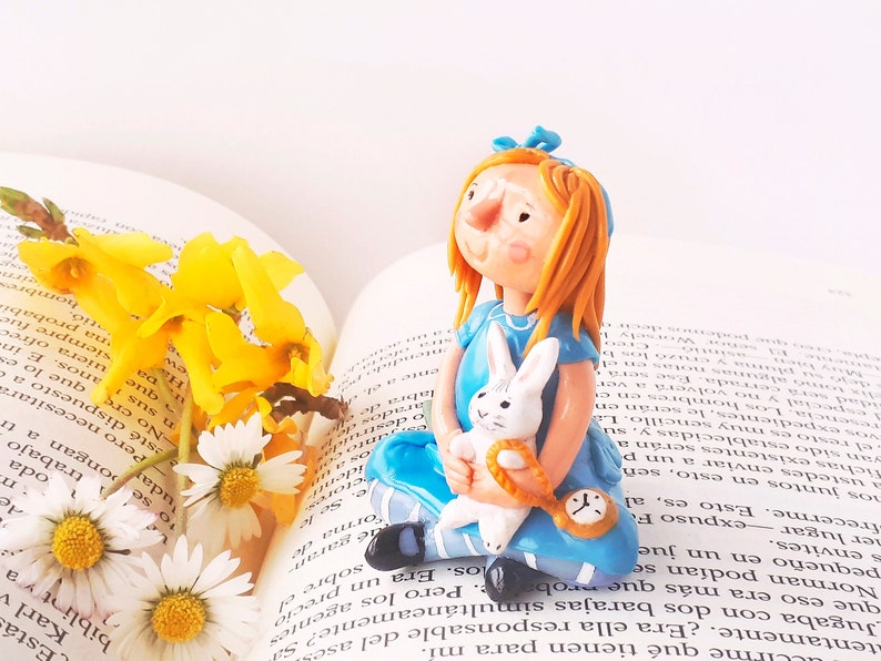 May include: A blue and white porcelain figurine of Alice from Alice in Wonderland holding a white rabbit. The figurine is sitting on an open book with yellow flowers.