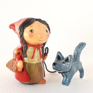 May include: A handmade figurine of Little Red Riding Hood with a red hooded cloak, brown hair, and a basket. She is walking with a blue wolf figurine.