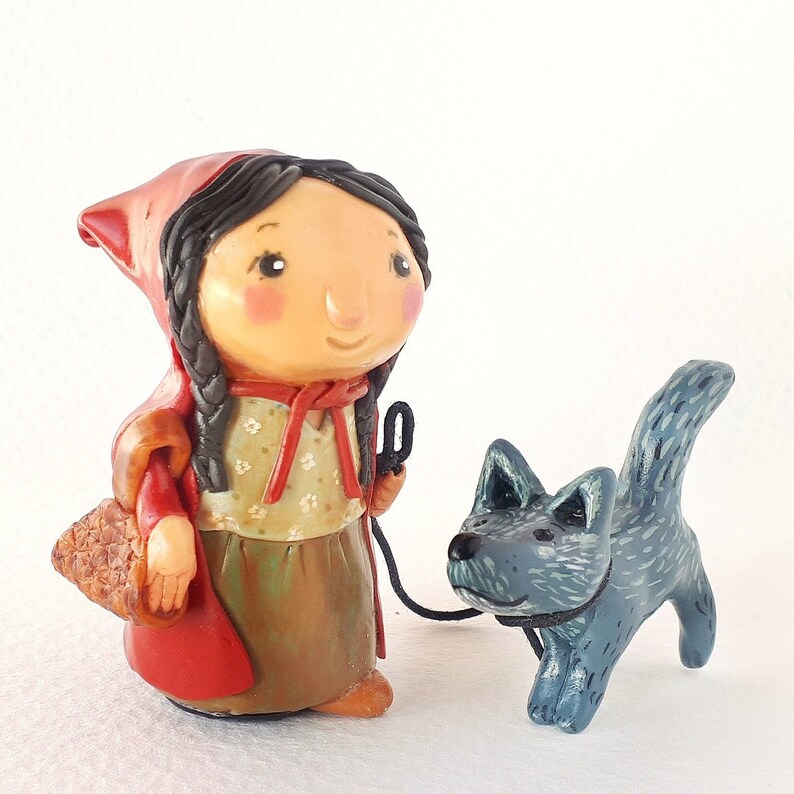 May include: A handmade figurine of Little Red Riding Hood with a red hooded cloak, brown hair, and a basket. She is walking with a blue wolf figurine.