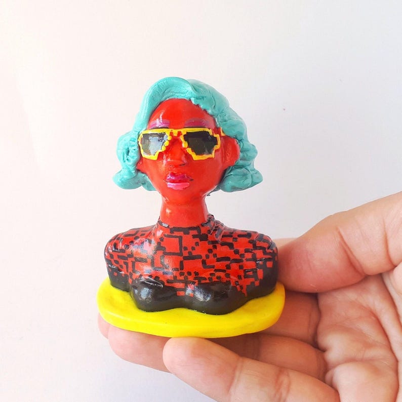 May include: A handmade ceramic bust of a person with blue hair, wearing red and black patterned clothing and yellow sunglasses.
