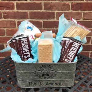 Może przedstawiać: Galwanizowany metalowy kubeł wypełniony zestawem do robienia s'mores. Kubeł ma nadrukowany tekst "Making More Memories" i "The De Simones". Zestaw zawiera krakersy graham, tabliczki czekolady, pianki i drewniane szaszłyki.