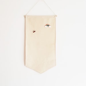 Peut inclure: Une bannière en tissu beige avec une épingle en forme d'arc-en-ciel et une épingle en forme de voiture rouge. La bannière est accrochée à un mur blanc.