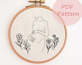May include: A round embroidery hoop with a linen fabric featuring a minimalist line drawing of a pregnant person. Black floral designs are at the bottom. The text "PDF Pattern" is in a pink circle.