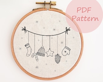 May include: A black and white embroidery pattern of a Christmas garland with a stocking, hat, star, pine cone, and mitten hanging from it. The pattern is on a white fabric background in a wooden embroidery hoop.