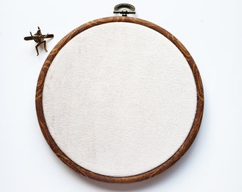 May include: A round wooden embroidery hoop with a white fabric center, featuring a bronze-colored clasp and a small decorative figure. The wood has a visible grain pattern, and the fabric is tightly stretched.