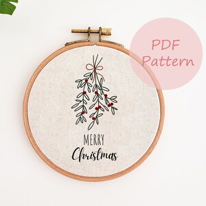 Könnte beinhalten: Ein runder Stickrahmen mit einem Leinenstoff mit einem schwarzen und roten Mistelzweig-Design und den Worten "MERRY Christmas" in schwarzer Schrift. Der Rahmen ist aus Holz und hat einen Messingverschluss. Ein rosa Kreis mit den Worten "PDF Pattern" befindet sich im Hintergrund.