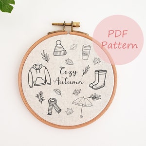 May include: A round embroidery hoop with a linen fabric featuring black line drawings of autumn-themed items. The design includes a hoodie, hat, coffee cup, leaves, boots, scarf, and umbrella. The words "Cozy Autumn" are written in cursive.